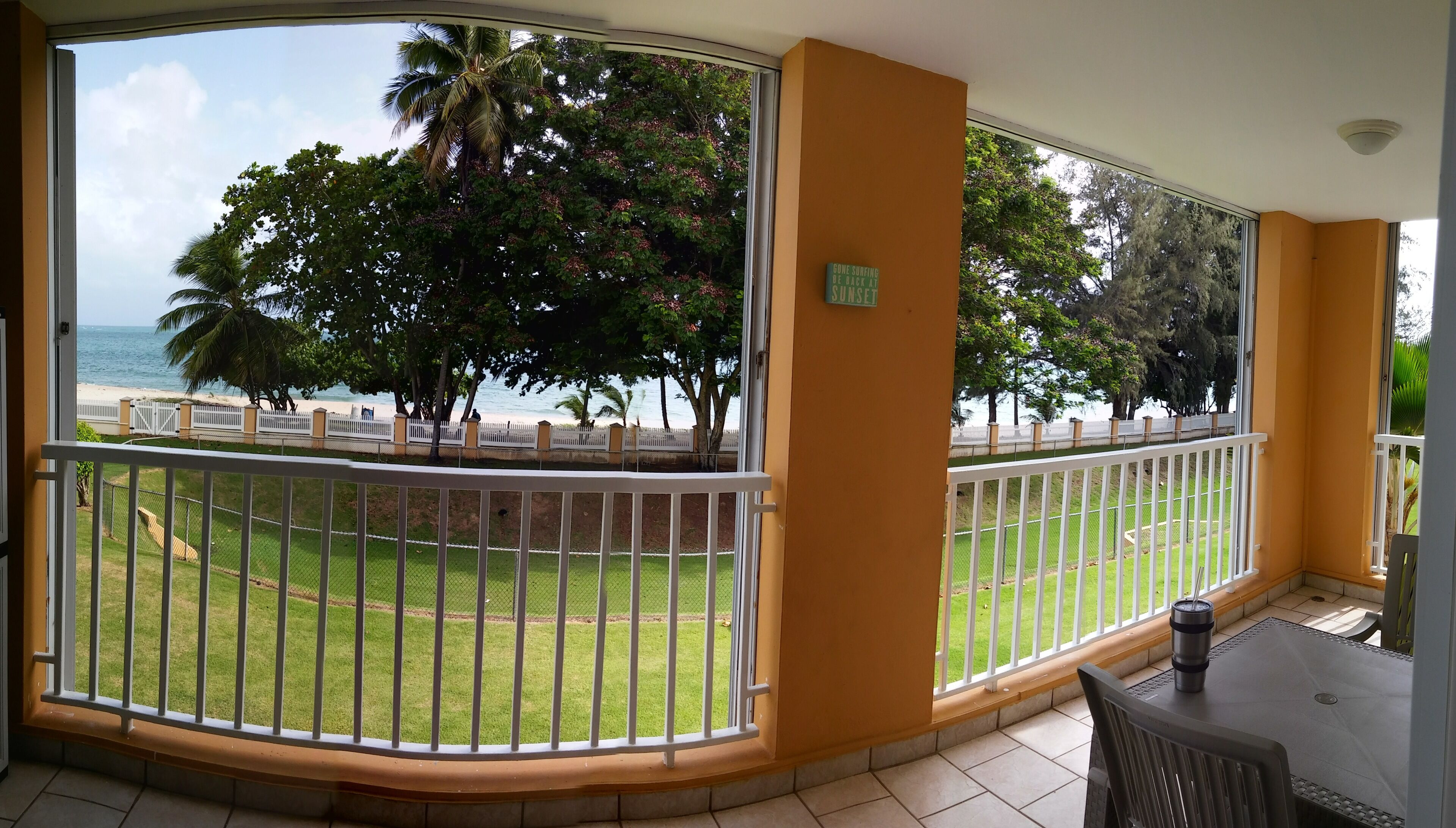 A balcony with a view of a beach, a fenced lawn, and a sign indicating a bone surfing beach.