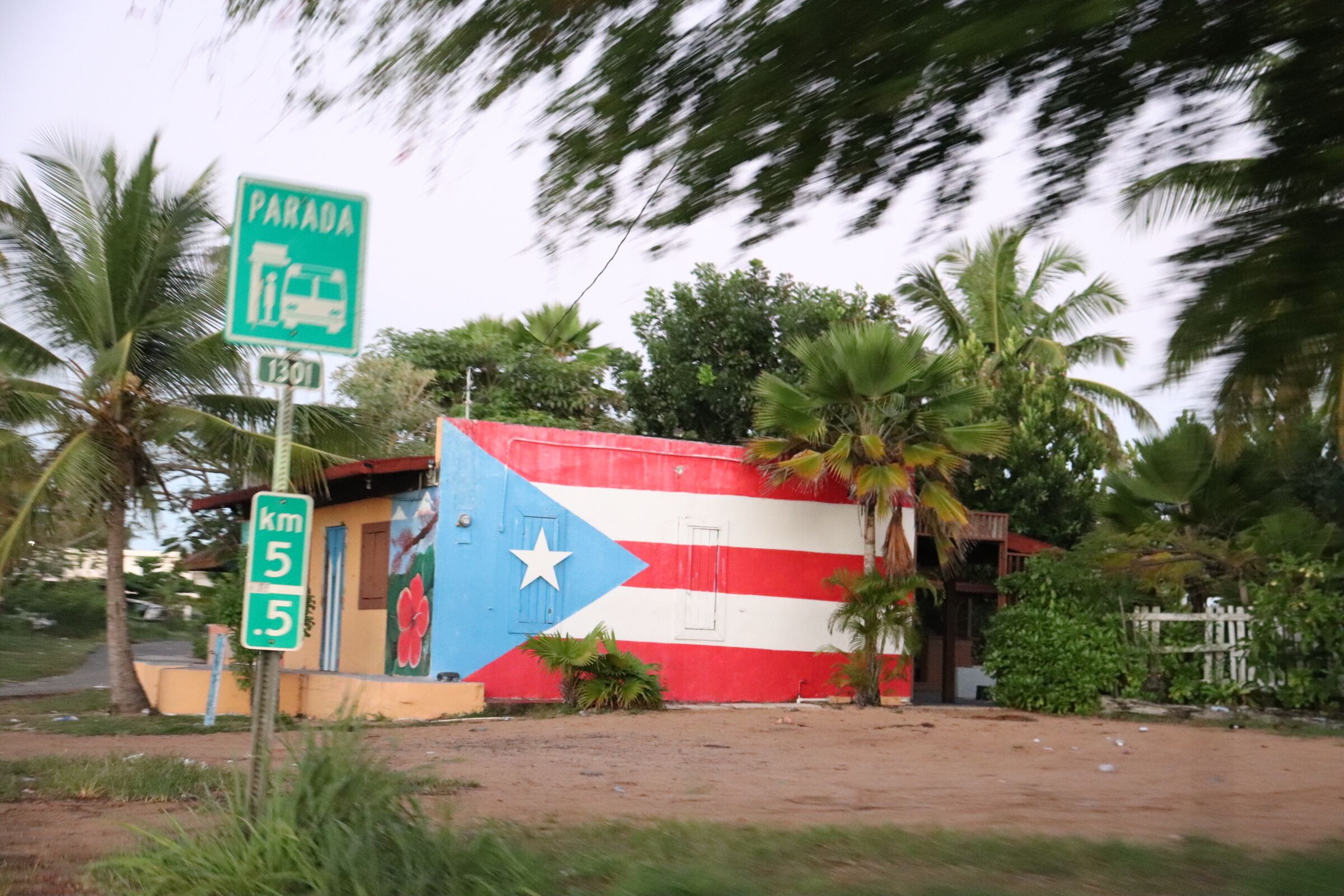 A building with a mural of a flag, a sign indicating 'Parada km 5.5', and palm trees in the background.
