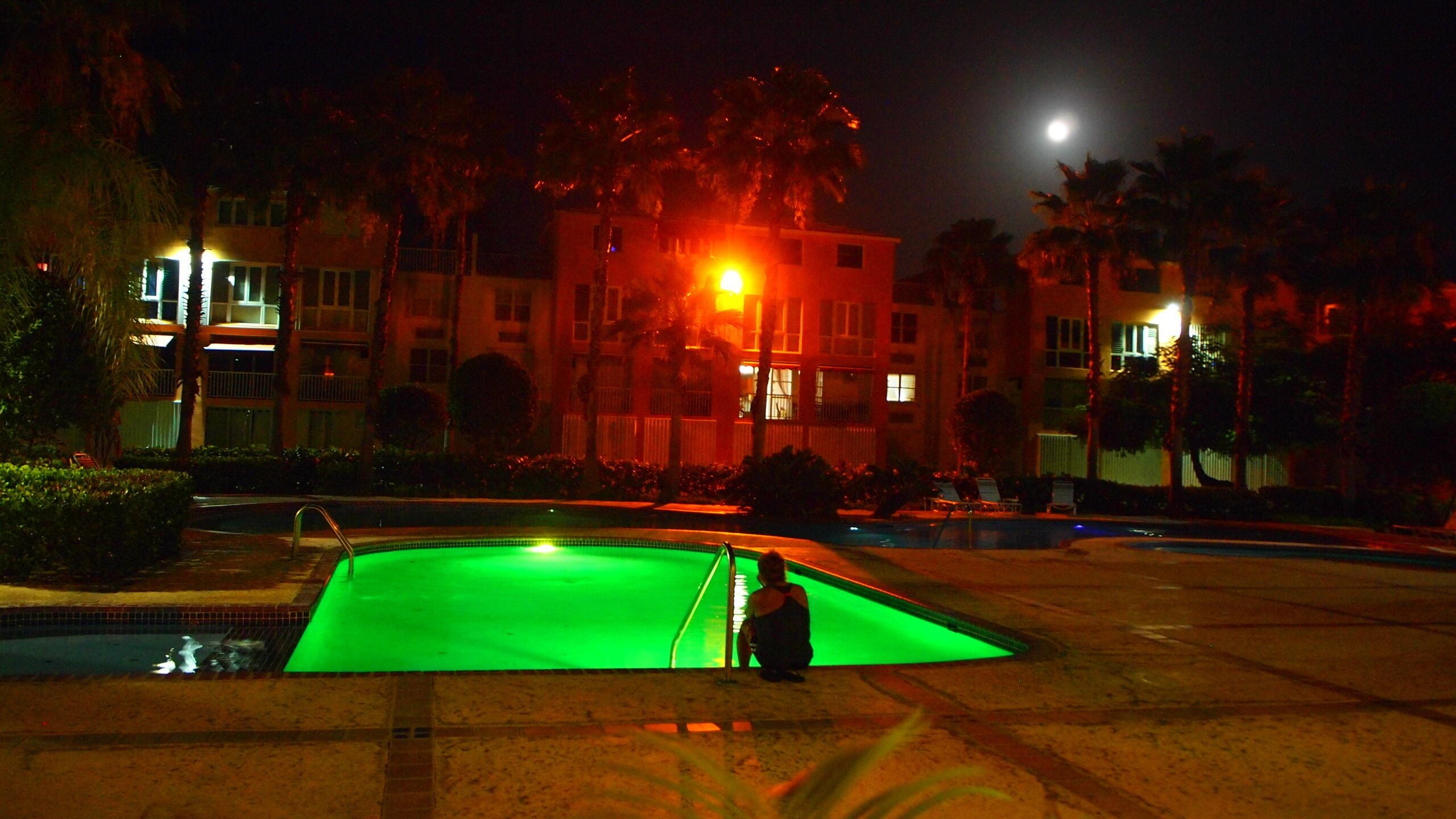 A nighttime pool area with a green-lit water feature, surrounded by palm trees and buildings.