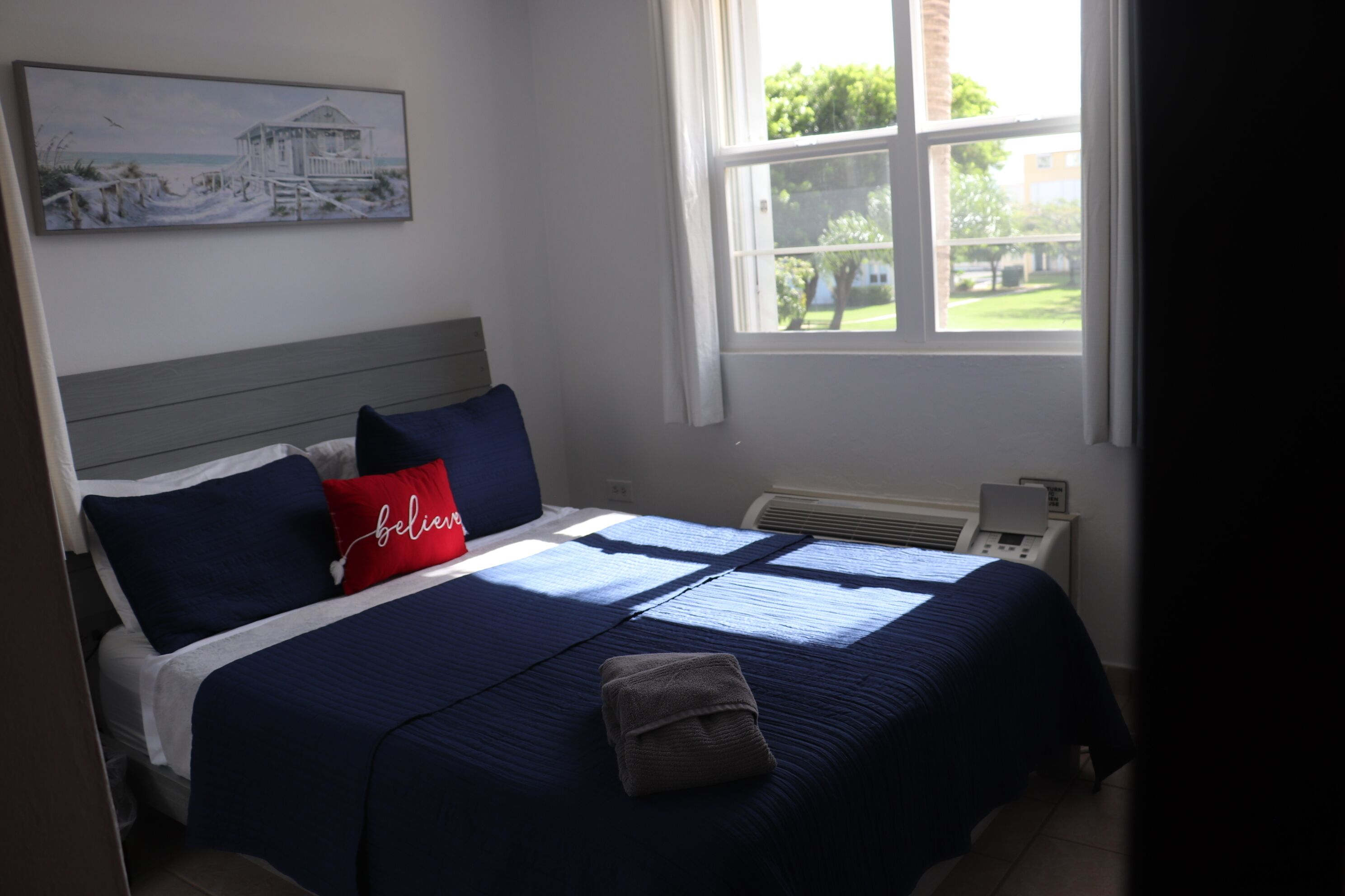 A neatly made bed with a blue bedspread, a red pillow with the word "believe," and a folded towel on the bed.