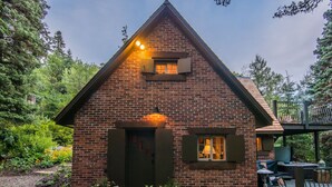 A brick cabin with a wooden deck, a small porch, and a window with shutters. - Wee Cottage- Unparalleled Charm, Tiny House, Enchanting Setting, Hot Tub (Sundance)