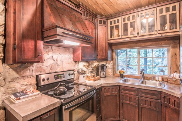 A rustic kitchen with stone walls, wooden cabinets, a stove, and a window with a view of trees. - Wee Cottage- Unparalleled Charm, Tiny House, Enchanting Setting, Hot Tub (Sundance)