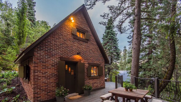 A rustic cabin with a brick exterior, a wooden deck, and a small porch area. - Wee Cottage- Unparalleled Charm, Tiny House, Enchanting Setting, Hot Tub (Sundance)