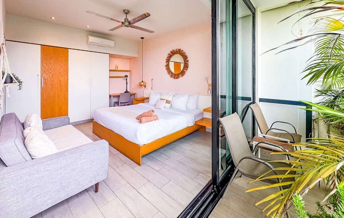 A modern hotel room with a bed, a sofa, a desk, and a balcony with plants.