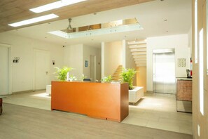 A modern reception area with a wooden reception desk, potted plants, a staircase, and a refrigerator.
