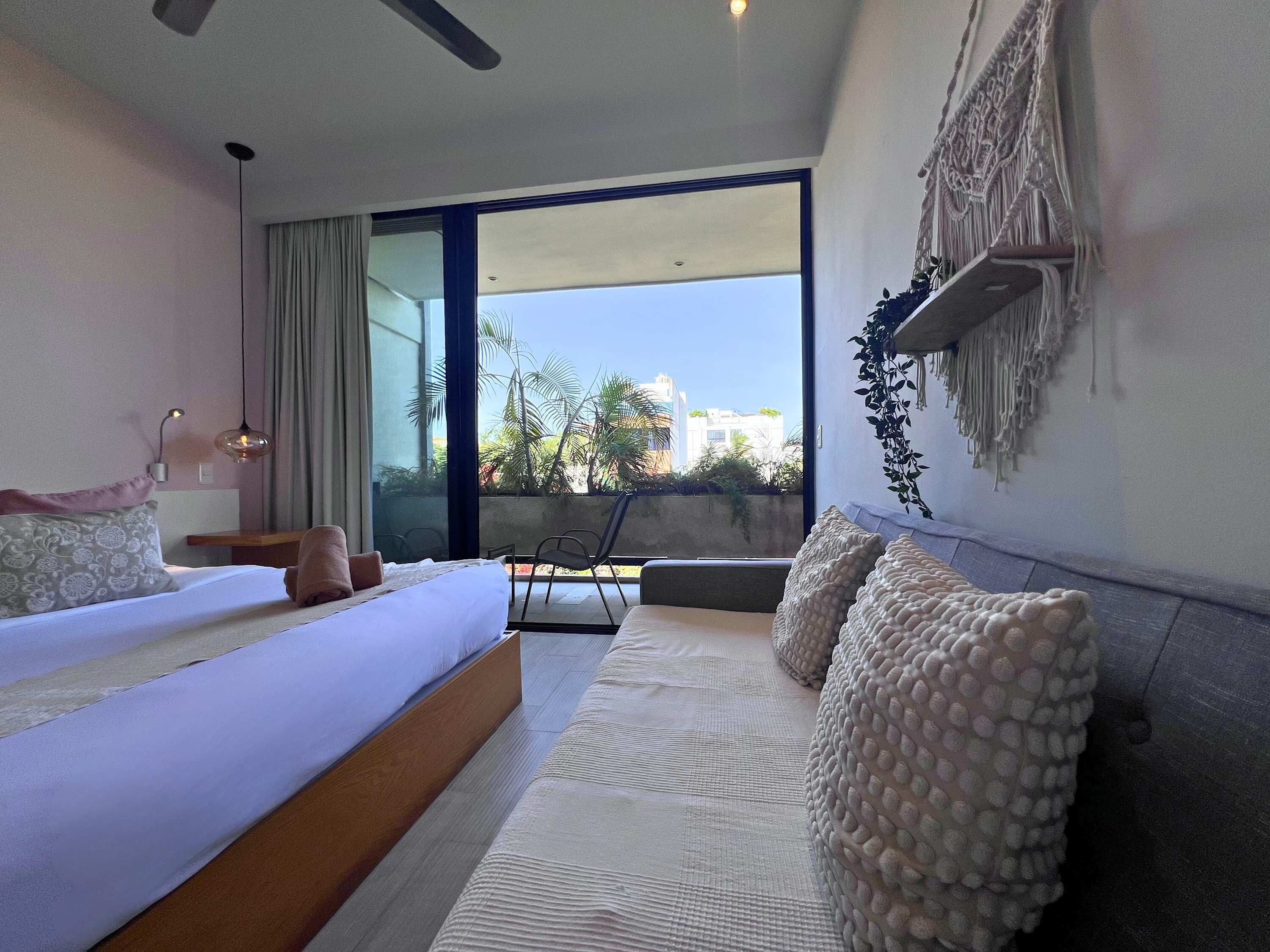 A modern bedroom with a large bed, a sofa, and a balcony with a view of buildings and trees.
