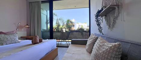 A modern bedroom with a large bed, a sofa, and a balcony with a view of buildings and trees.