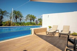 A poolside area with a clear blue pool, two lounge chairs, and a white wall.