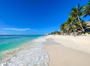Ein Strand mit klarem türkisblauem Wasser, weißem Sand und Palmen. Entlang der Küste befinden sich Liegestühle und Sonnenschirme.