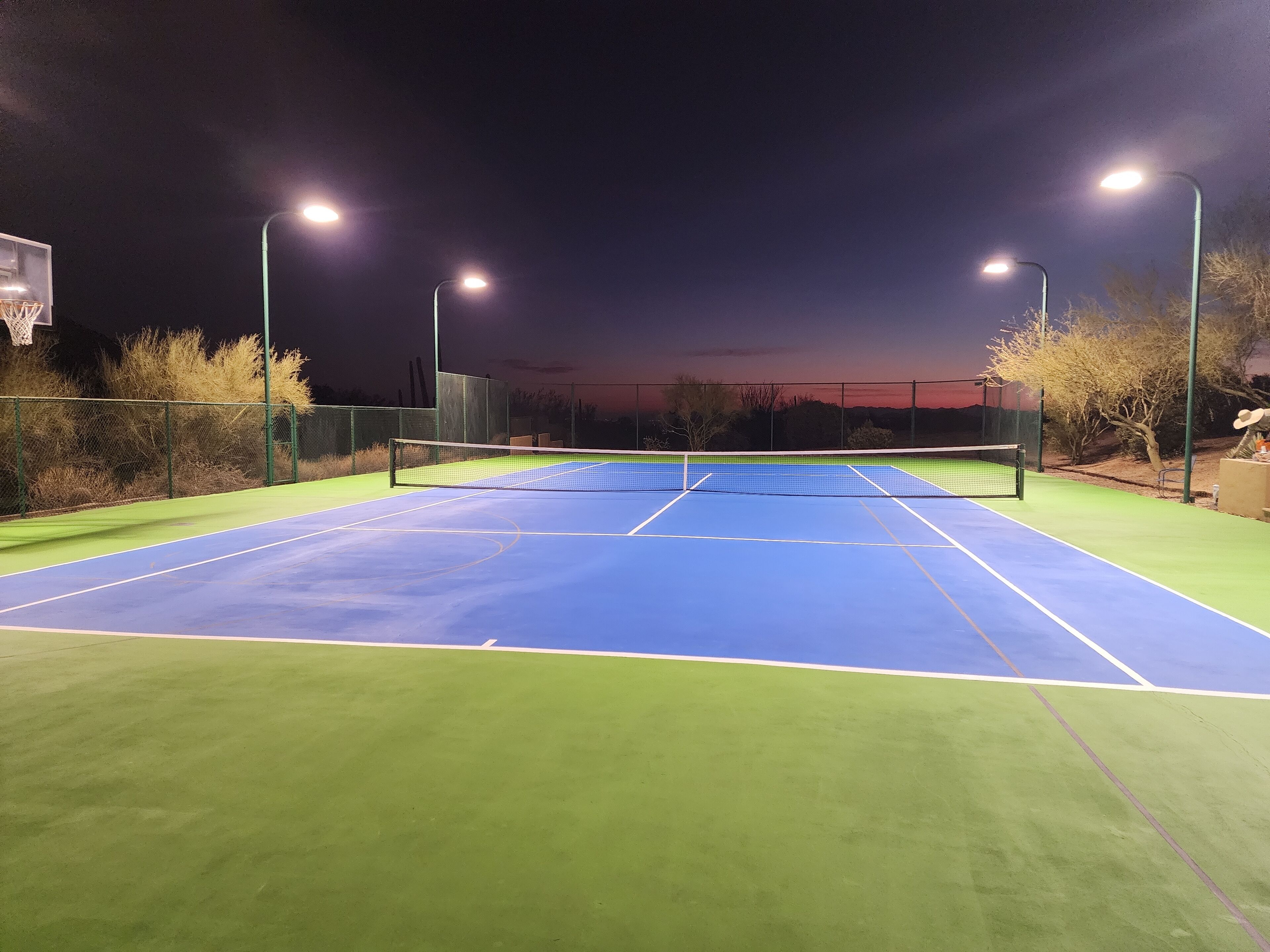 Un court de tennis extérieur, éclairé par des projecteurs la nuit, entouré d’une clôture et d’arbres.
