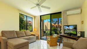 A living room with a sofa, a television, and a balcony with a view of palm trees.