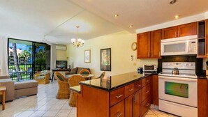 A kitchen with wooden cabinets, a microwave, and an oven. There is a dining area with wicker furniture and a view of palm trees through a glass door.