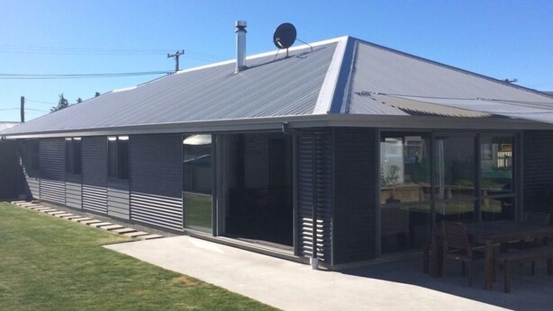 A modern building with a metal roof, large glass windows, and a covered outdoor area with furniture.
