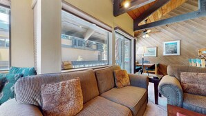 A cozy living area with a sofa, a table, and a chair. There is a window with a view of a balcony and a ceiling fan. - Secluded Cozy Cabin, Forest Setting, 1-Car Garage, Near Winter Shuttle (Mammoth Lakes)