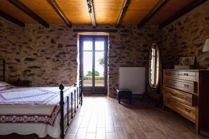 A bedroom with a stone wall, wooden floor, a bed with a white and purple bedspread, a wooden dresser, and a large window with a view outside. - Mystras Village House (Sparta)