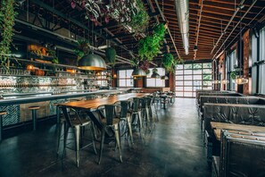 Un grand espace bar avec de hauts plafonds, des poutres apparentes et des plantes suspendues. On y trouve plusieurs coins salon avec des tabourets et des tables.