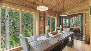Un coin repas rustique en bois, avec un banc et une table, de grandes fenêtres donnant sur la forêt, et un intérieur chaleureux agrémenté de poutres en bois et de luminaires suspendus.