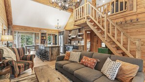 Un intérieur de chalet chaleureux avec un escalier en bois, un canapé agrémenté de coussins à motifs, un coin repas et une cuisine.