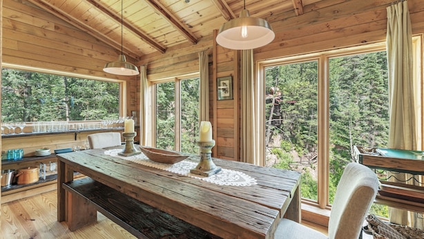 Un intérieur rustique en bois, avec une grande table à manger, des bougies et une vue sur une forêt à travers de grandes fenêtres.