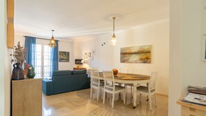 Uma área de refeições com uma mesa redonda, cadeiras brancas e um candelabro. Há um sofá e um armário ao fundo.