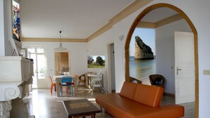 Una sala de estar con un sofá, una mesa de comedor con sillas, una mesa de centro de madera y vista a una costa rocosa a través de un arco.