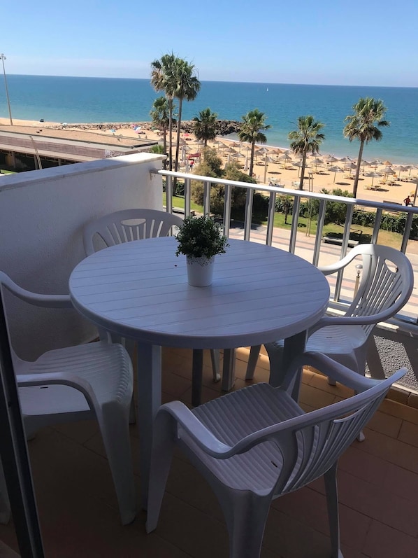 Un balcon avec une table ronde et des chaises en plastique blanc, donnant sur une plage bordée de palmiers et surplombant un ciel bleu azur.