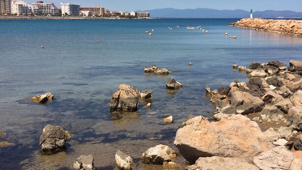 Escena costera con aguas cristalinas, costa rocosa y un paisaje urbano distante.