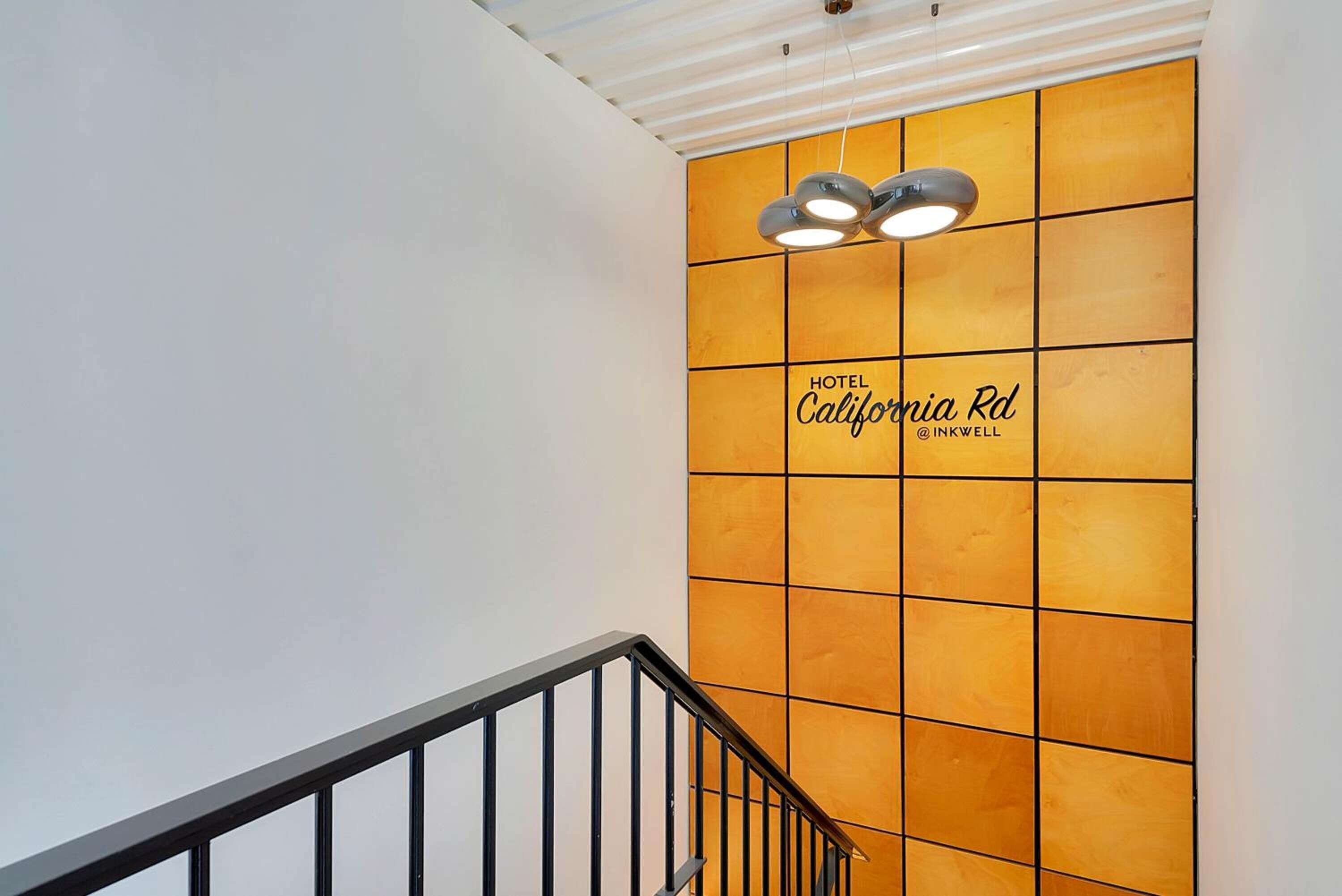 Eine Treppe mit Metallgeländer führt zu einer Wand mit einem Raster aus orangefarbenen Tafeln und einem Schild, auf dem steht: HOTEL California Rd @ INKWELL.
