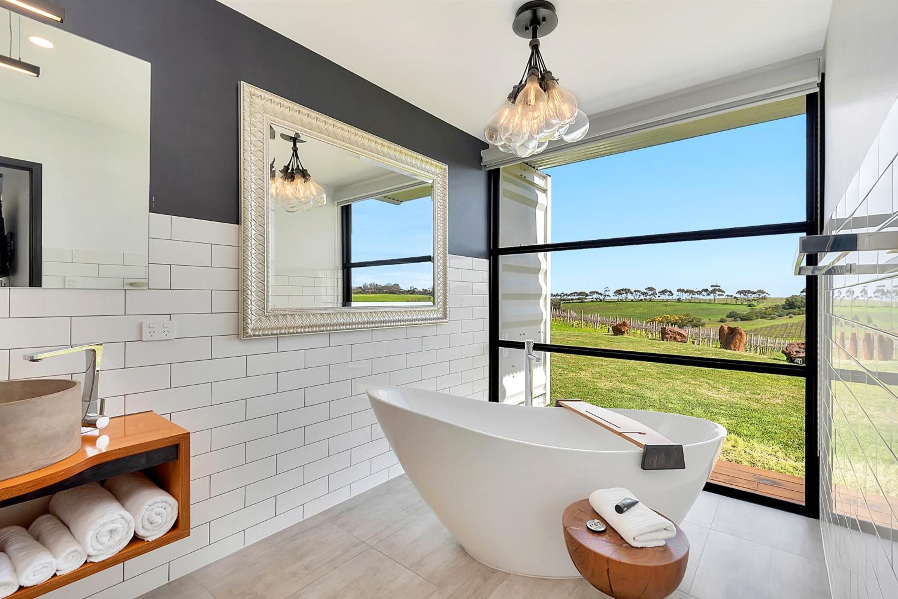 A modern bathroom with a freestanding bathtub, a large mirror, and a view of a vineyard through large windows.
