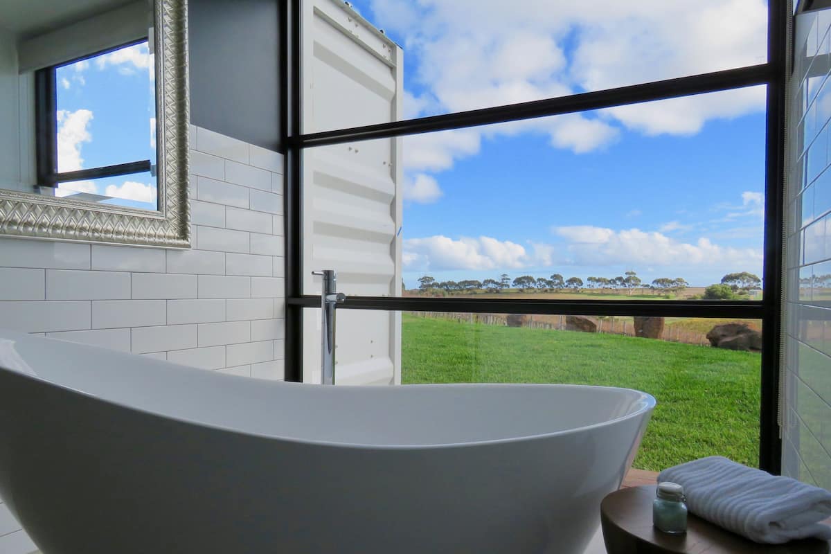A modern bathroom with a freestanding bathtub, a small table with a towel and bottle, and a large window offering a view of a green field and trees.