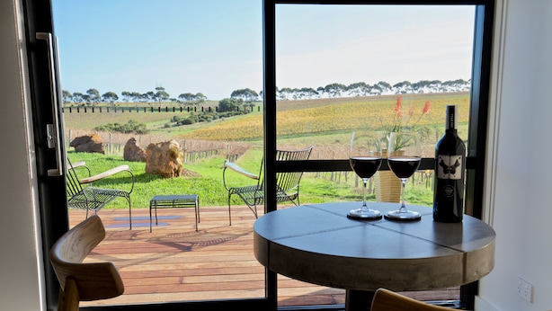 A room with a large window overlooking a vineyard, a round table with two wine glasses and a bottle, and a green cushioned chair. - Hotel California Road at Inkwell Wines (Tatachilla)