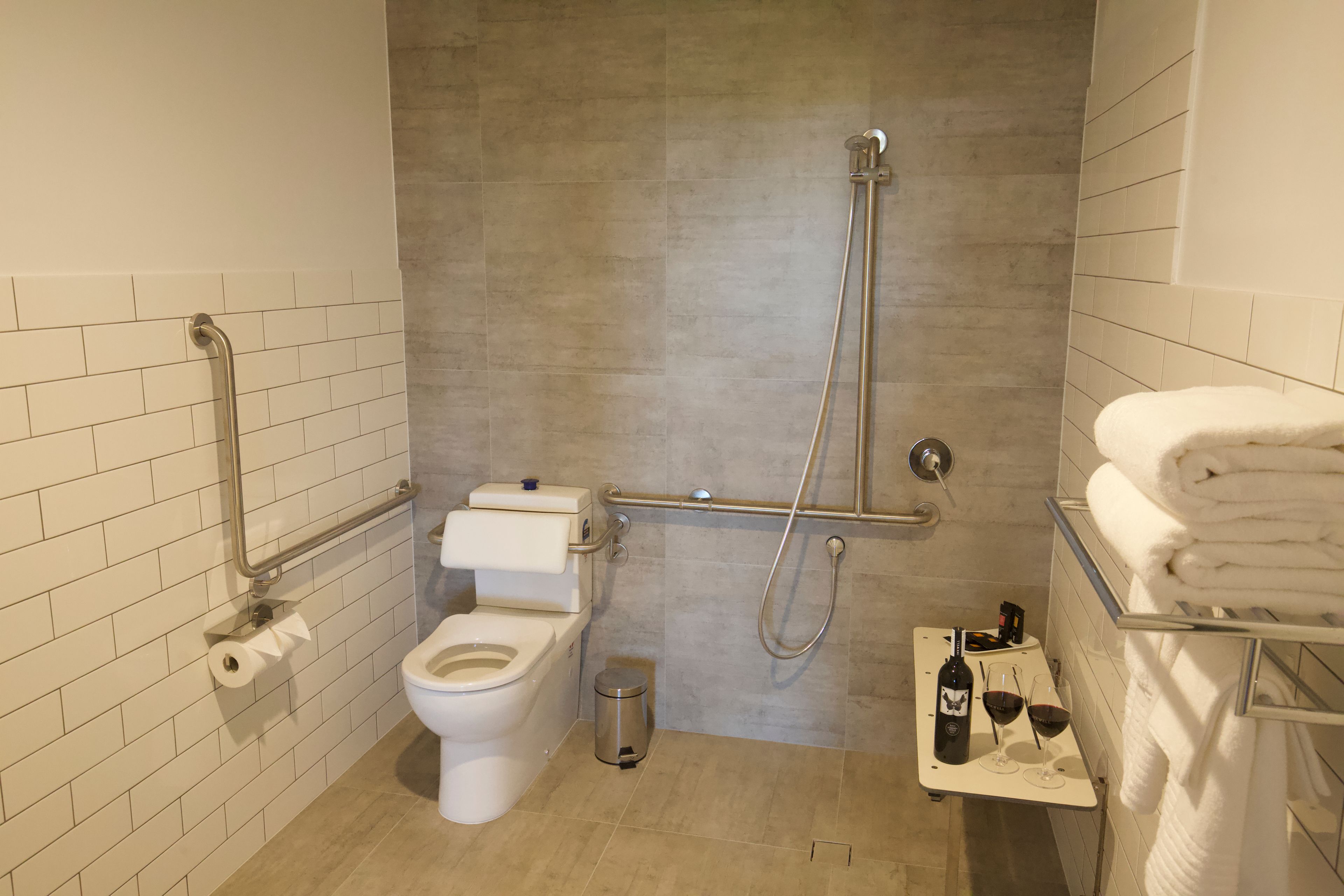 Un baño moderno con inodoro, bidé y ducha. Hay un cubo de basura y un estante con toallas y una botella de vino.