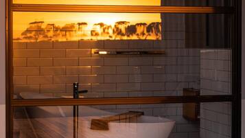 Un baño moderno con una bañera independiente, cabina de vidrio y vistas a un paisaje urbano al atardecer.