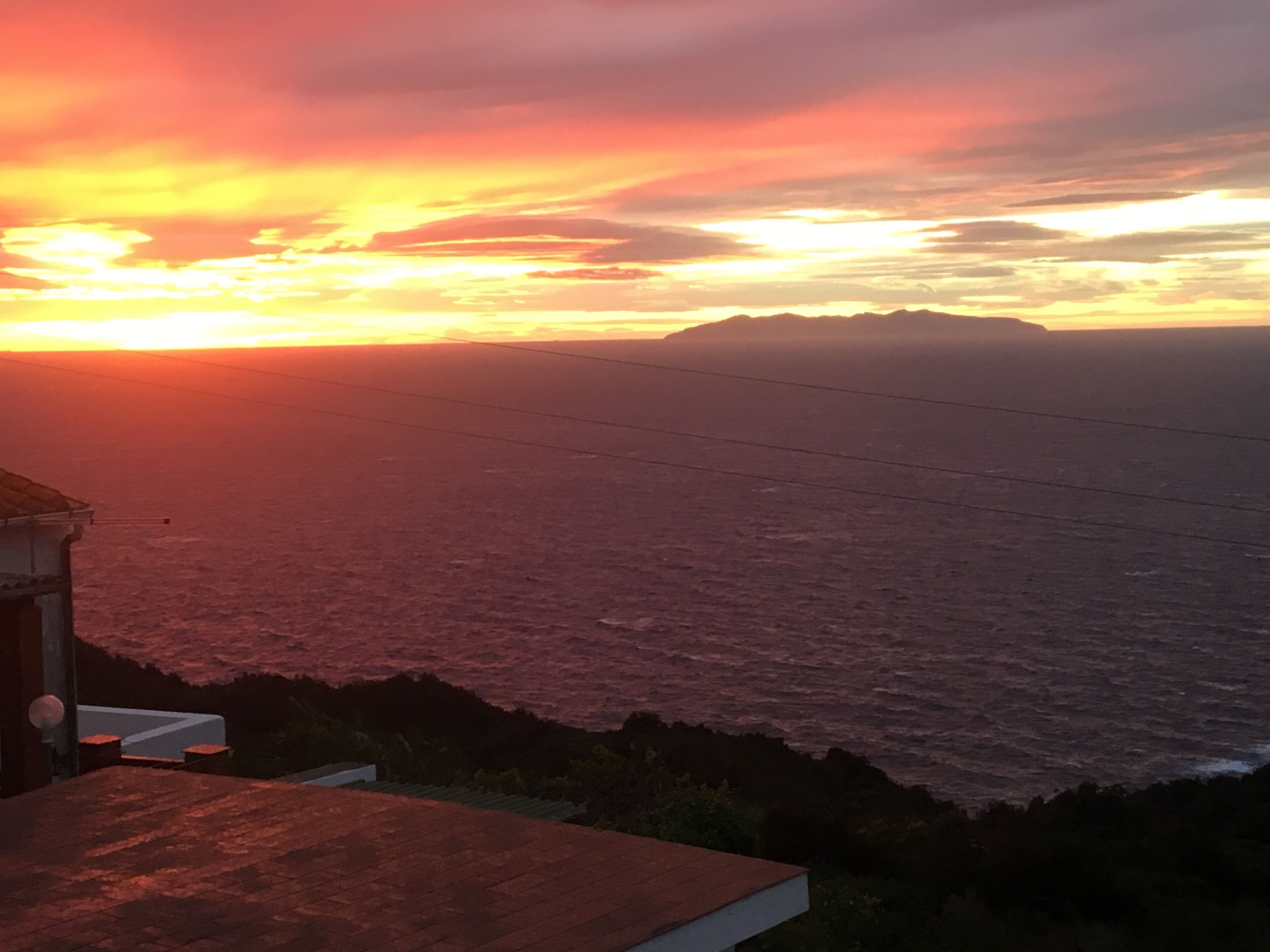 Ein Küstenbild mit Sonnenuntergang, ein Gebäude mit geneigtem Dach und eine entfernte Bergkette.