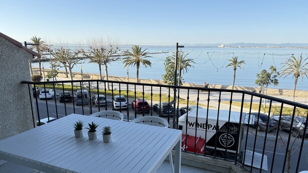Un balcon avec une table et des chaises blanches, donnant sur un parking où sont garées des voitures et sur une plage.