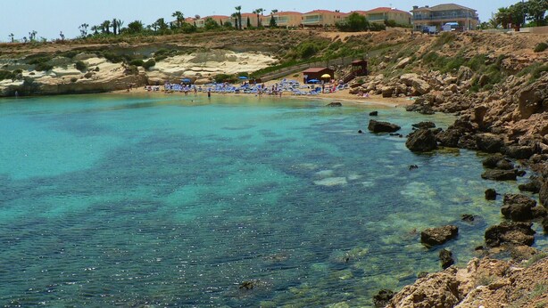 Eine Küstenlandschaft mit klarem türkisblauem Wasser, felsigem Ufer und einer Reihe von Strandgebäuden mit Sonnenschirmen.