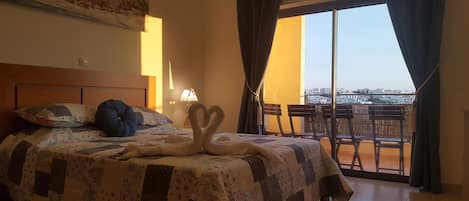 A hotel room with a bed, a painting on the wall, a view of the city through the window, and a balcony with chairs.