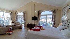 Une chambre d’hôtel avec un grand lit, un canapé, un fauteuil, une télévision et un balcon donnant sur des immeubles.