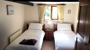 Una habitación con dos camas individuales, una mesita de noche con lámpara, una ventana con cortinas y una puerta.