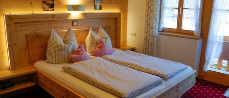 Ein Bett mit Holzbettgestell, karierter Bettwäsche und Kissen, ein Nachttisch und ein Fenster mit Vorhängen.
