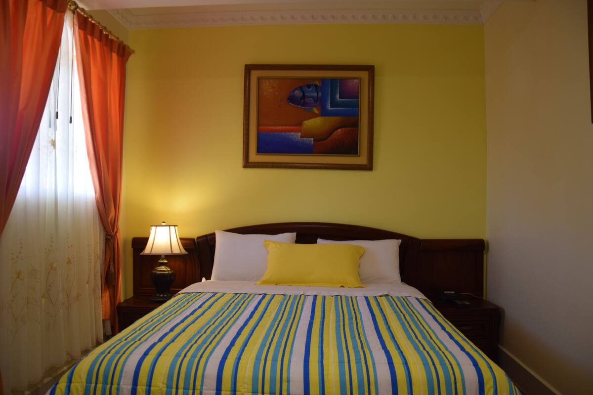 Ein Hotelzimmer mit Bett, Nachttisch, Lampe, einem Bild an der Wand und Vorhängen.