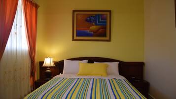 Quarto de hotel com cama, mesa de cabeceira, abajur, pintura na parede e cortinas.