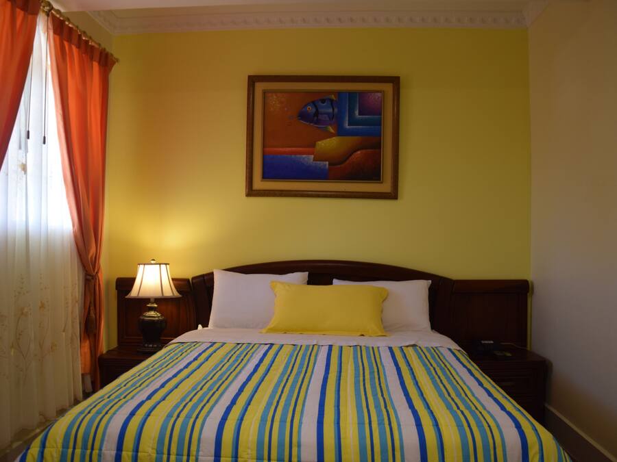 Quarto de hotel com cama, mesa de cabeceira, abajur, pintura na parede e cortinas.