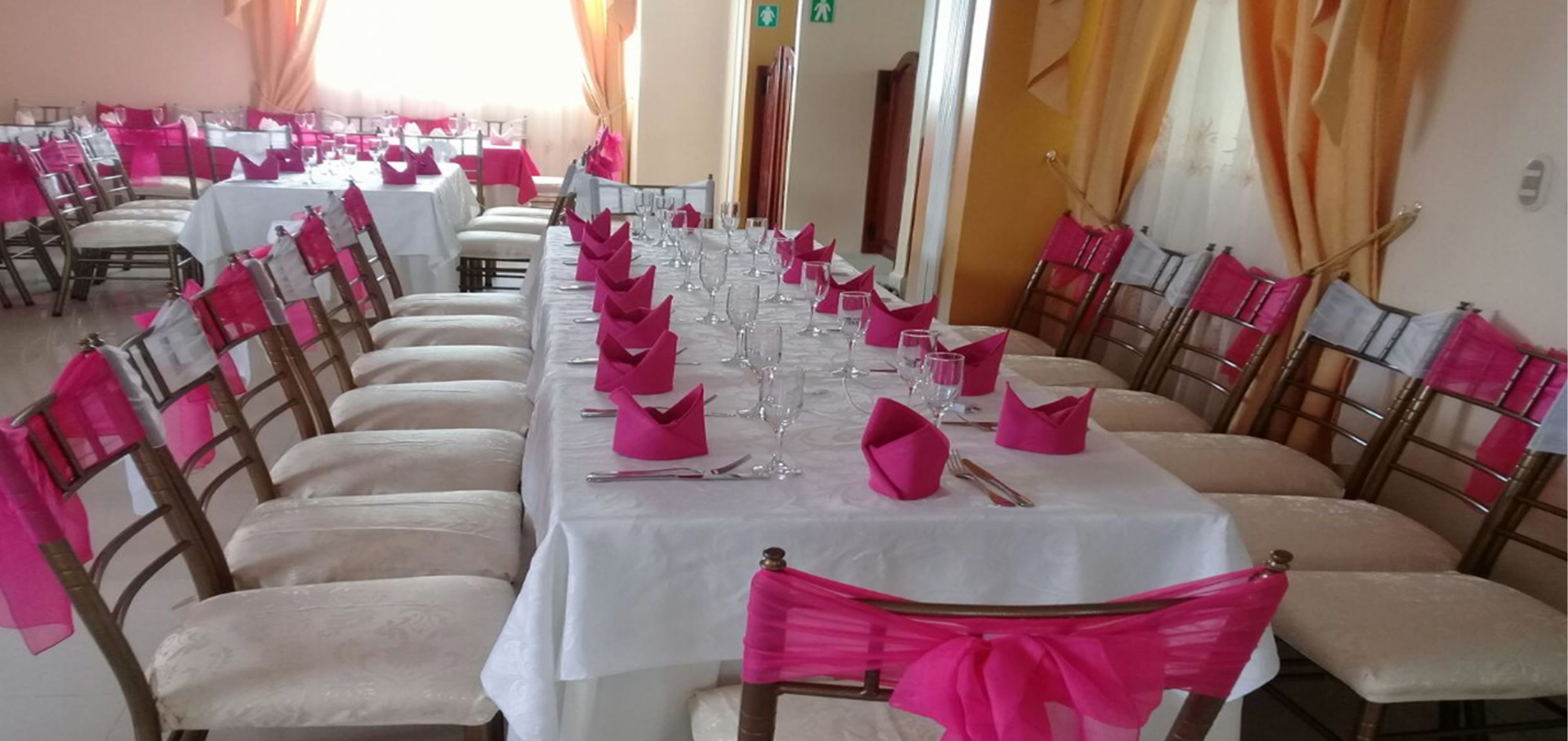 Una larga mesa de comedor dispuesta con servilletas rosas, cubiertos y copas de vino, rodeada de sillas adornadas con cintas rosas.