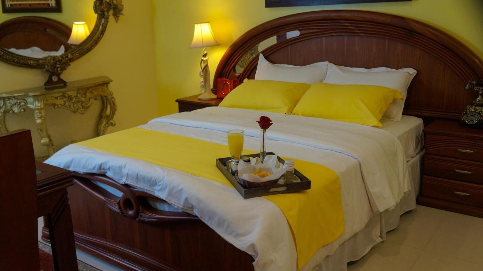 Una cama bien hecha con una bandeja de desayuno, que incluye un vaso de jugo de naranja y una rosa.