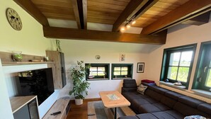 A modern living room with a sofa, a coffee table, and a dining area. There are two windows with green frames, a plant, and a clock on the wall. - Vacation home Hanse in the vacation village Altes Land - Holiday home 327 Hanse 80sqm for max. 7 persons (Hollern-Twielenfleth)