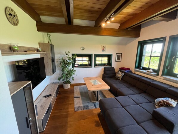 A modern living room with a sofa, a coffee table, and a dining area. There are two windows with green frames, a plant, and a clock on the wall.