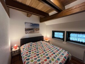 A bedroom with a bed, two bedside lamps, a window with curtains, a picture on the wall, and a wooden ceiling.