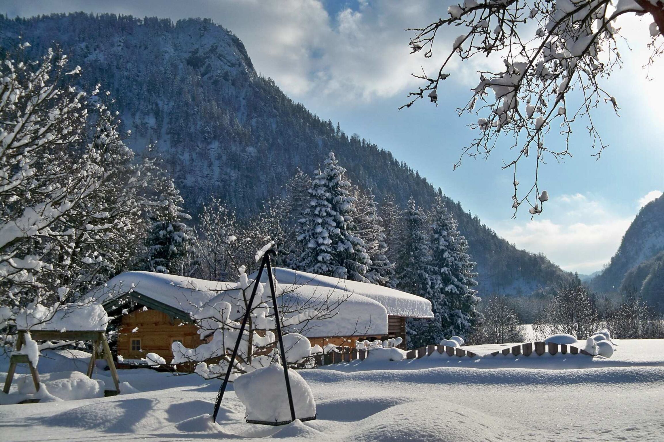 Eine Holzhütte mit schneebedecktem Dach, umgeben von schneebedeckten Bäumen und Bergen im Hintergrund.