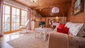 L’intérieur d’un chalet en bois chaleureux et accueillant, avec une grande fenêtre, un fauteuil à bascule, un canapé agrémenté de coussins à motifs floraux et un lustre.